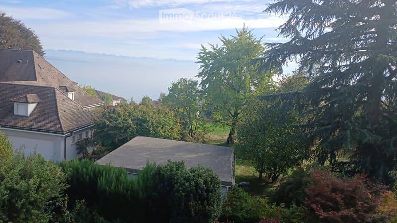 A two-story house with a tiled roof and white exterior, surrounded by lush greenery and trees, with a scenic view of a lake and distant mountains.