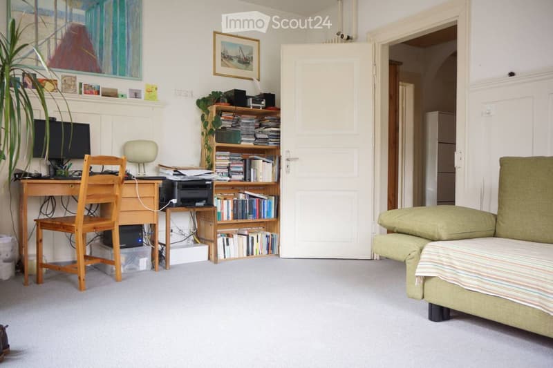 A cozy, well-furnished room with a sofa, desk, chair, lamp, printer, bookshelf, screen, table, and plant. The room features a large open doorway leading to another space.