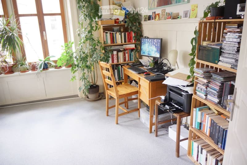 A well-lit study or home office with a wooden desk, computer setup, bookshelf filled with books, and several potted plants near the window.