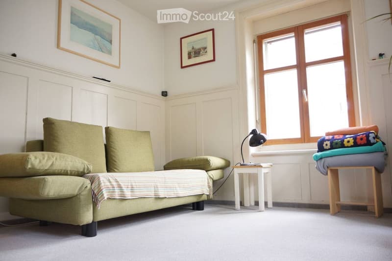 A cozy living room with a green sofa, small white table, black lamp, and a wooden stool with folded blankets.