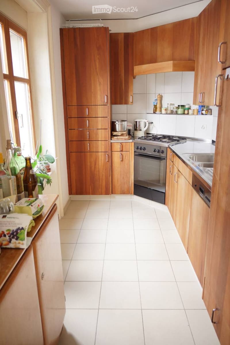 A modern kitchen with wooden cabinets, white tiled floor, and stainless steel appliances including a stove, oven, and sink. There are various kitchen items on the counter and a small plant near the window.