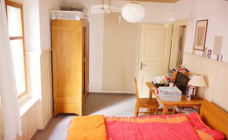 Cozy bedroom with a bed, wardrobe, desk, chair, lamp, and a mirror. The room features a window with white curtains and a door leading to another room.
