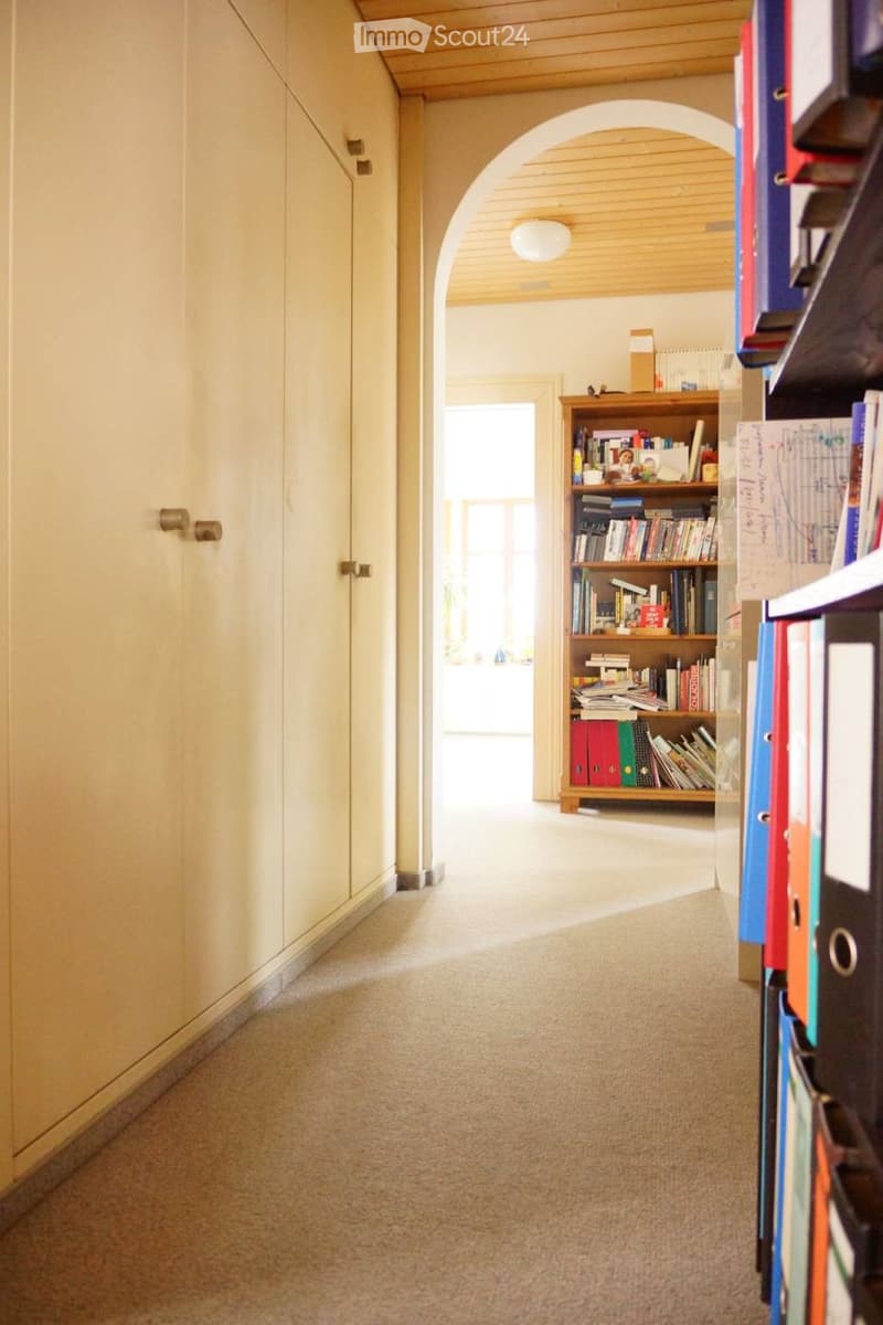 Carpeted hallway with light-colored walls, wooden ceiling, and a wooden bookshelf filled with books and binders on the right side.