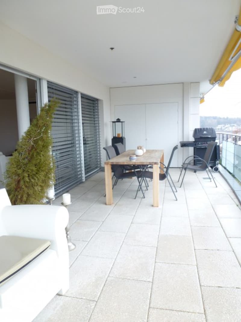 Modern balcony with tiled floor, large windows, dining table with chairs, grill, and a small tree.