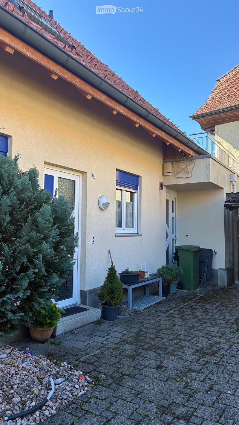Outdoor patio area with a small table, potted plants, and a trash bin, adjacent to a house with a tiled roof and windows.