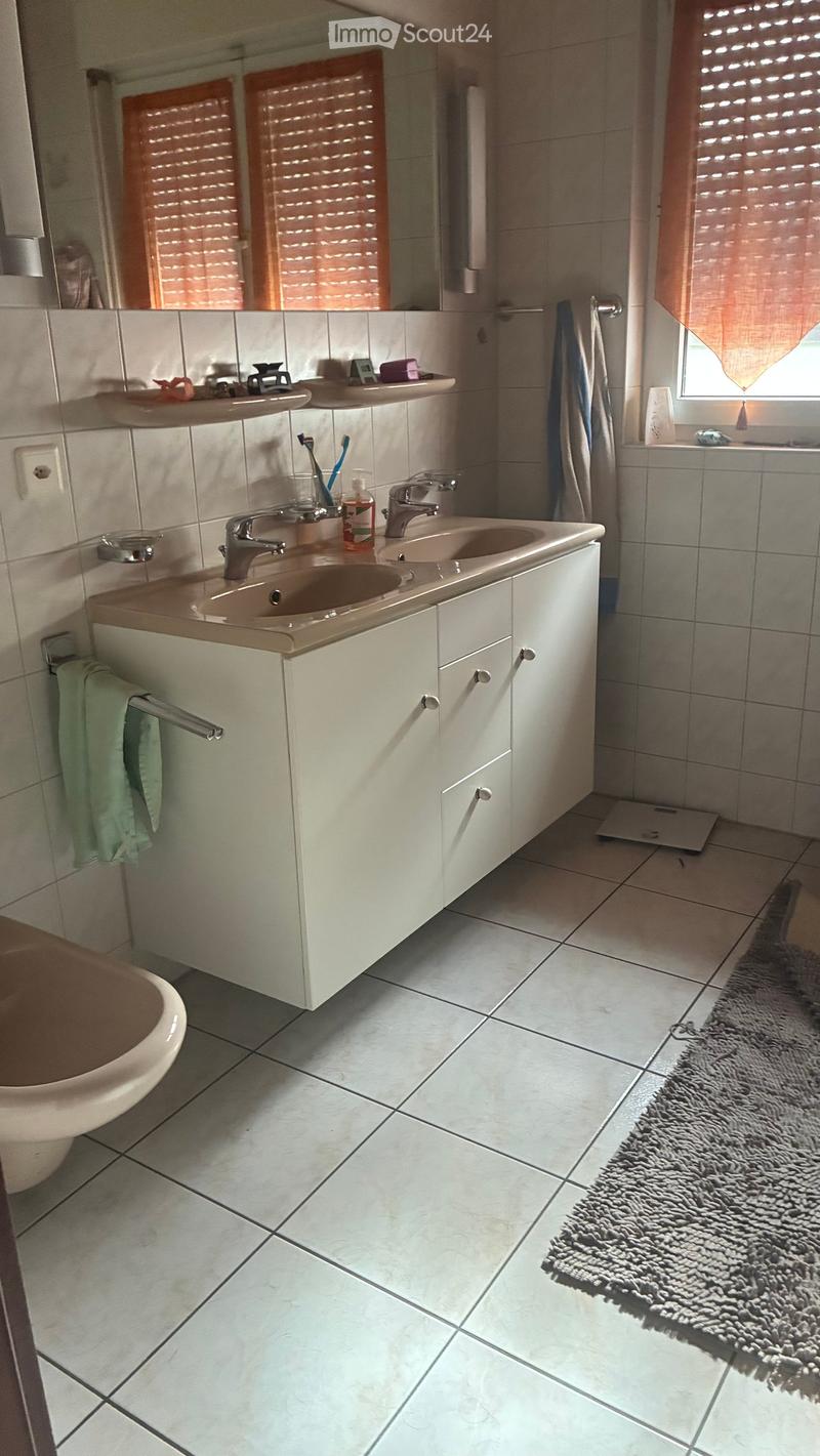 Modern bathroom with white tiles, double sink, mirror, shelves, and a bathtub. Includes a towel, toothbrush, toothpaste, and a rug on the floor.