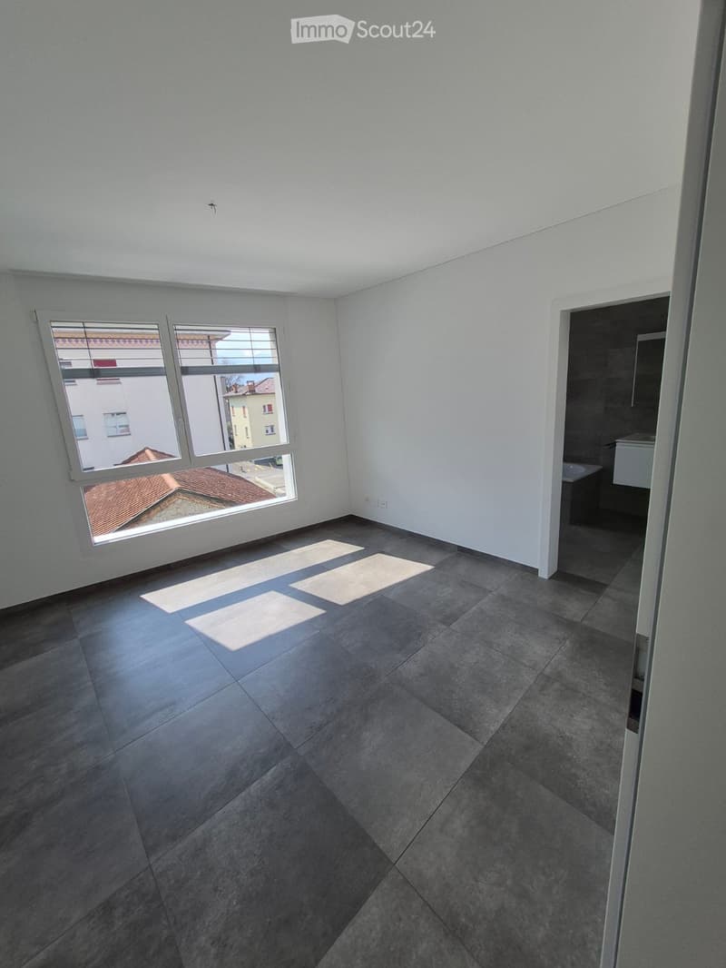Empty room with grey tiled floor, white walls, and large windows showing a view of neighboring buildings. Sunlight streams in, casting shadows on the floor.