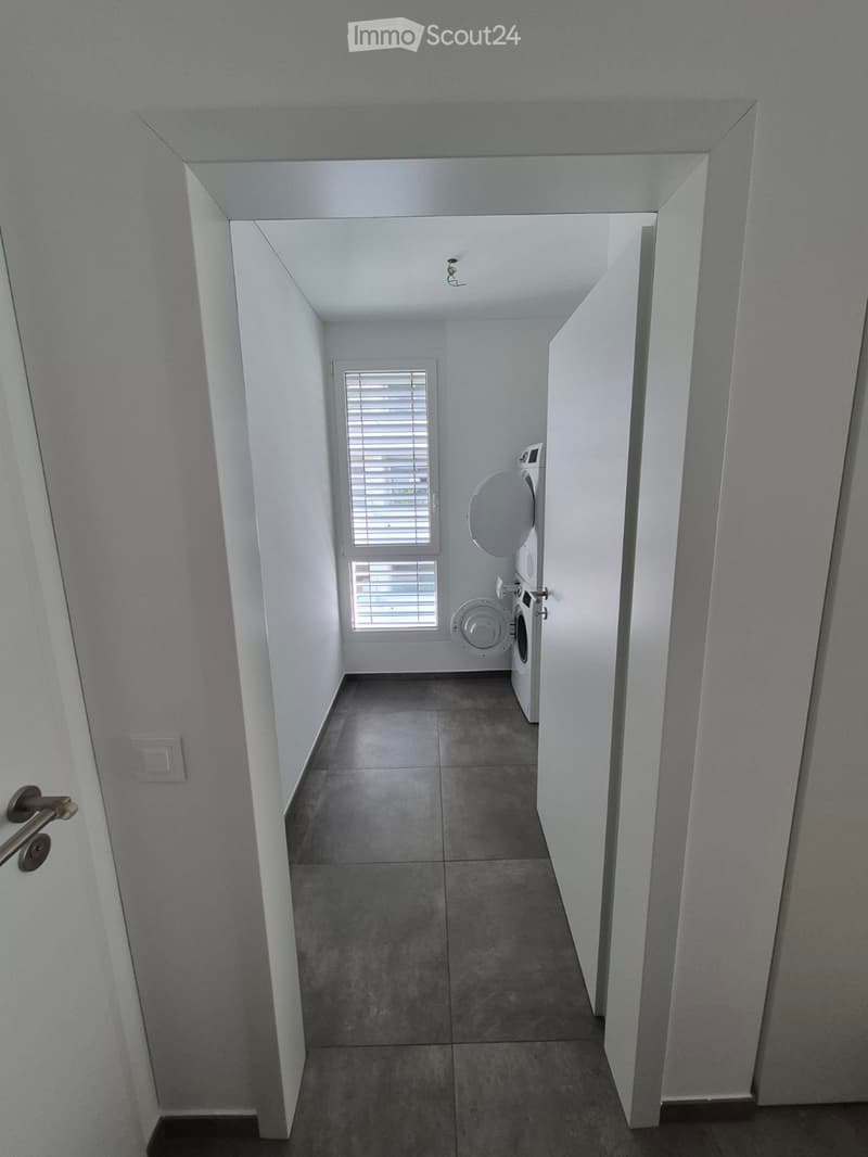 A narrow, well-lit hallway with tiled flooring and white walls, leading to a room with a washing machine and dryer visible at the end.