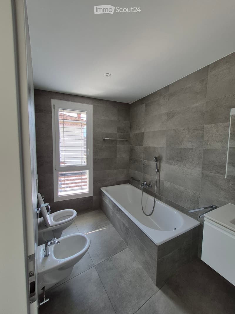 Modern bathroom with grey tiled walls and floor, featuring a bathtub, toilet, sink, and mirror. A window with blinds provides natural light.