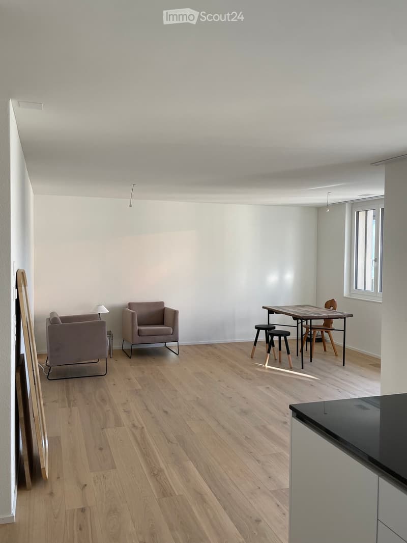 Bright, open living space with light wooden flooring, two armchairs, a small table with stools, and a lamp near the wall.