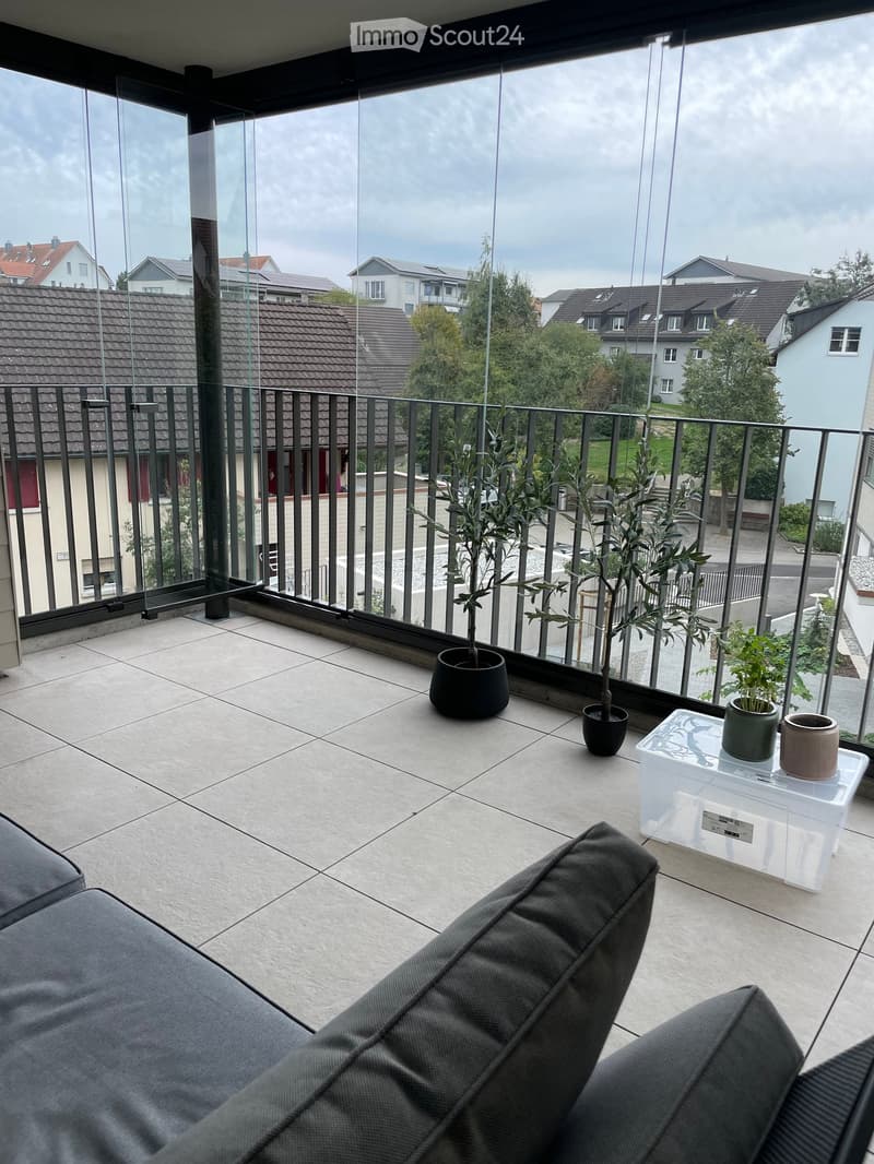 Balcony with tiled floor, glass railing, and view of surrounding houses. Features a grey sofa, small table with potted plants, and additional potted plants.