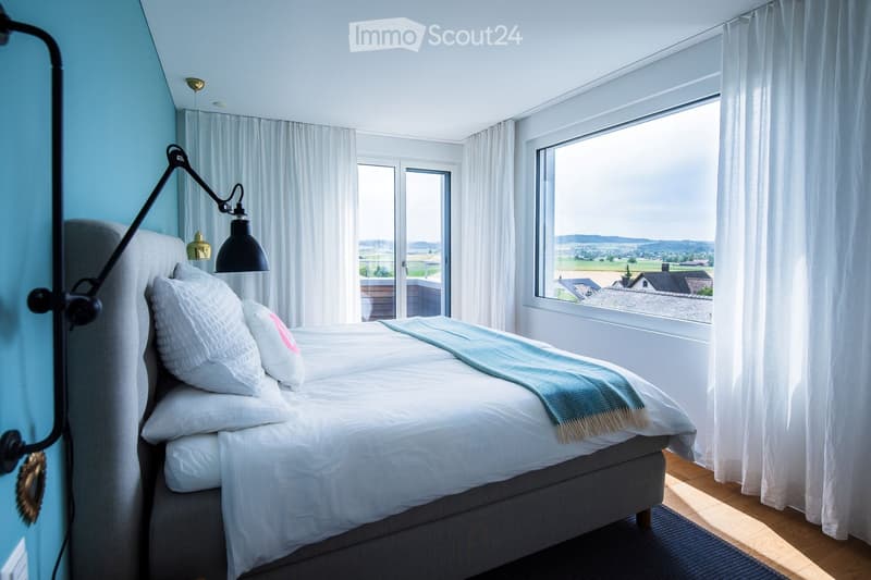 Bright bedroom with a large bed, pillows, a blanket, a wall-mounted lamp, and large windows offering a scenic view of the countryside.