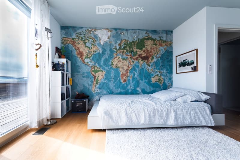 Modern bedroom with a large world map mural, a white bed with pillows, a nightstand, a bookshelf, a floor lamp, and a framed picture on the wall.