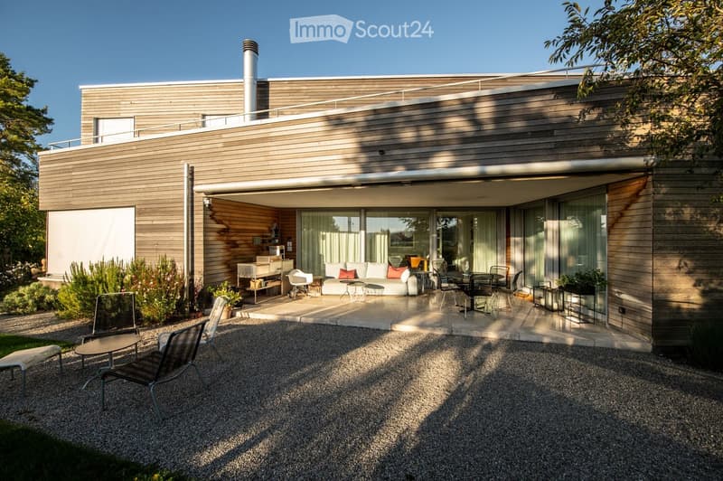 Modern house with a spacious outdoor patio area featuring a sofa, tables, and chairs. The patio is connected to the house via large glass doors, and the surrounding area includes gravel pathways and greenery.