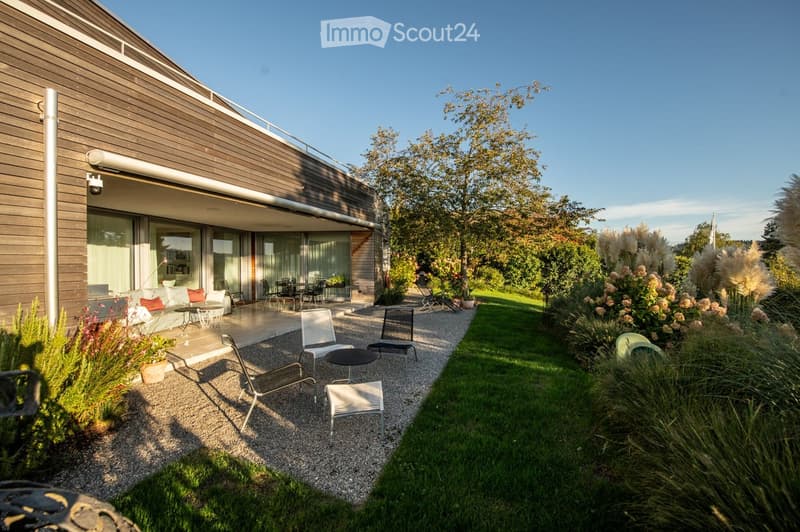 Modern outdoor patio area with a gravel pathway, lounge chairs, small table, and lush greenery including shrubs and tall grasses. The adjacent house features large glass windows and a wooden exterior.