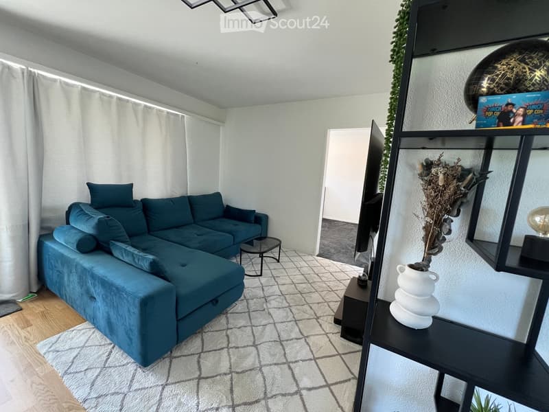 Modern living room with a blue sectional sofa, coffee table, shelving unit with decorative items, and a large screen. The space features a mix of wooden and tiled flooring, large windows with white curtains, and a cozy rug.