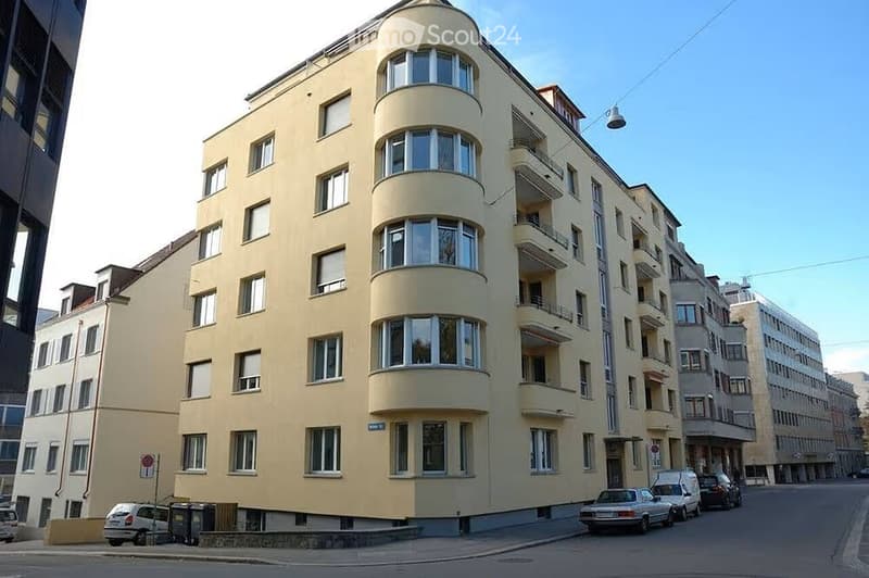 Multi-story residential building with light yellow facade, multiple windows, and balconies. Several cars are parked along the street in front of the building.