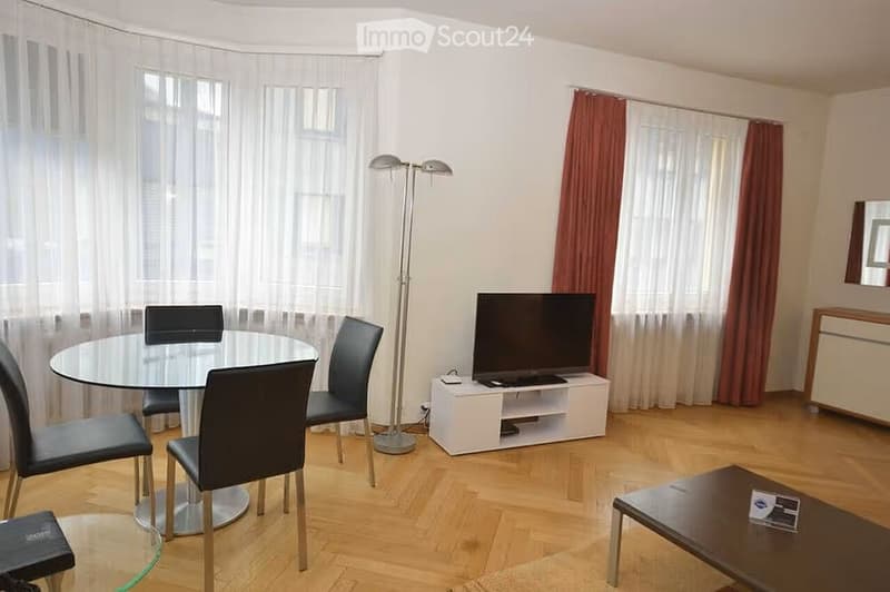 Modern living and dining area with wooden flooring, large windows with white and red curtains, a glass dining table with four black chairs, a flat-screen TV on a white stand, a coffee table, and a floor lamp.