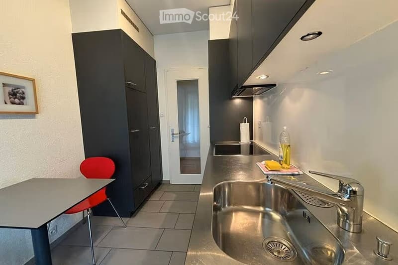 Modern kitchen with stainless steel sink, faucet, dark cabinets, and a small dining area with a table and red chair.