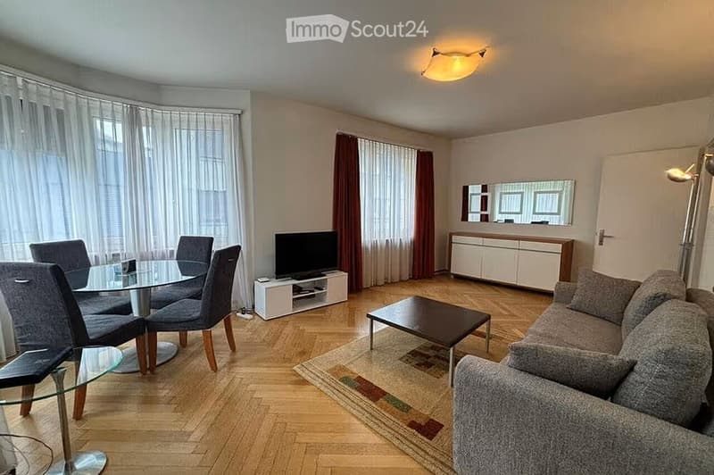 Modern living and dining area with a grey sofa, coffee table, dining table with chairs, TV, and a cabinet with a mirror above it. The space features wooden flooring, large windows with curtains, and a ceiling light fixture.