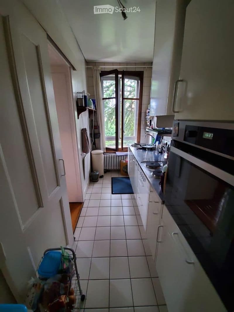 The kitchen features white tiled flooring, white cabinets, and a countertop with various appliances including an oven, microwave, and sink. There is a window with a view of greenery outside, and a small rug on the floor.