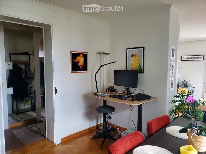 A modern, well-lit room with a wooden floor, a desk with a computer, a chair, a stool, a small table with chairs, and several picture frames on the walls.
