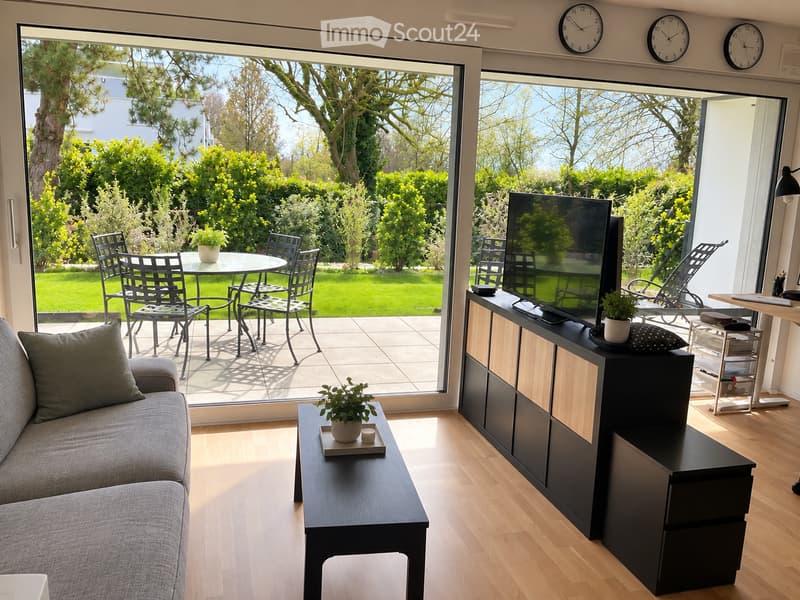 Bright living room with large windows and a view of a garden, featuring a grey sofa, coffee table, TV stand, and outdoor dining area with chairs and a table.