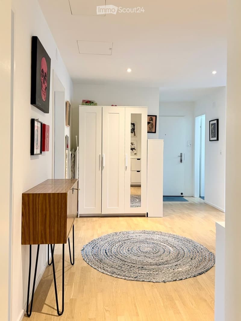 Modern hallway with light wooden flooring, white walls, a round woven rug, a wooden console table with metal legs, and a large white wardrobe with mirrored doors.