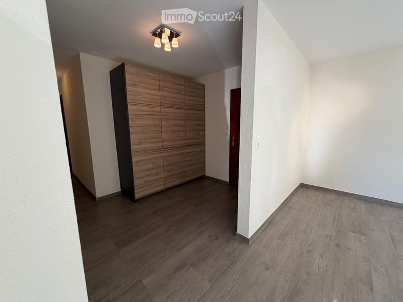 Empty room with wooden flooring, white walls, and a built-in wooden wardrobe. The room has a modern design with recessed lighting and a visible door leading to another area.