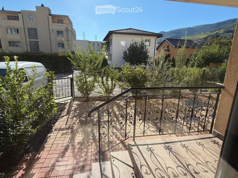 Balcony with wooden decking, metal railing, small table and chair, overlooking a residential area with trees and mountains.