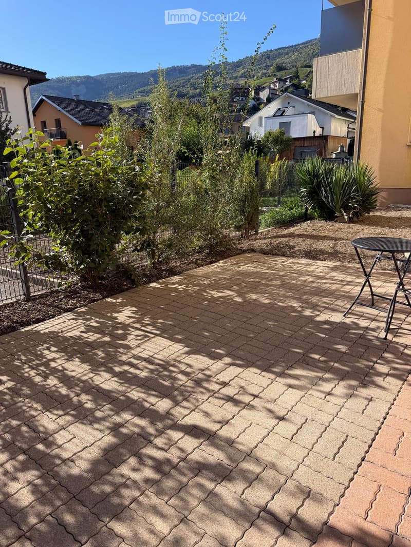 A well-maintained garden area with a paved patio, small table, and various plants, overlooking a scenic mountain landscape.