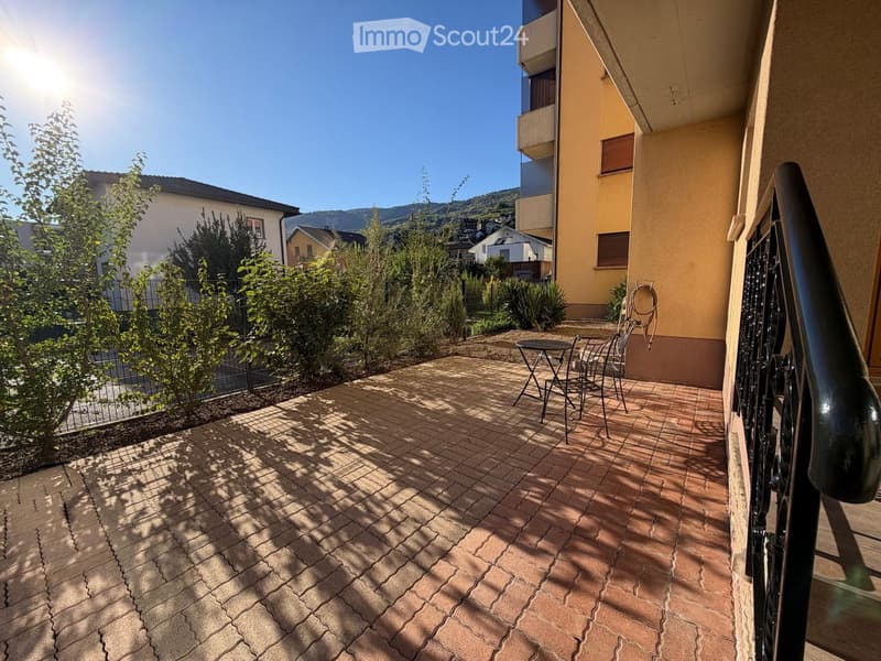 Balcony with brick tiles, small table and chairs, surrounded by greenery and a view of the town and mountains.