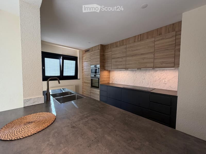 Modern kitchen with wooden cabinets, stainless steel sink, and built-in oven. Features a large window with blinds and a woven mat on the floor.
