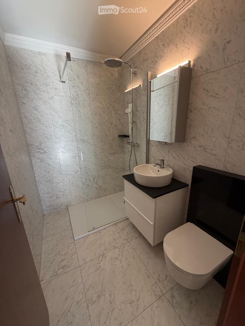 Modern bathroom with marble tiles, a glass-enclosed shower, a white sink with a modern faucet, a toilet, and a built-in mirrored cabinet.