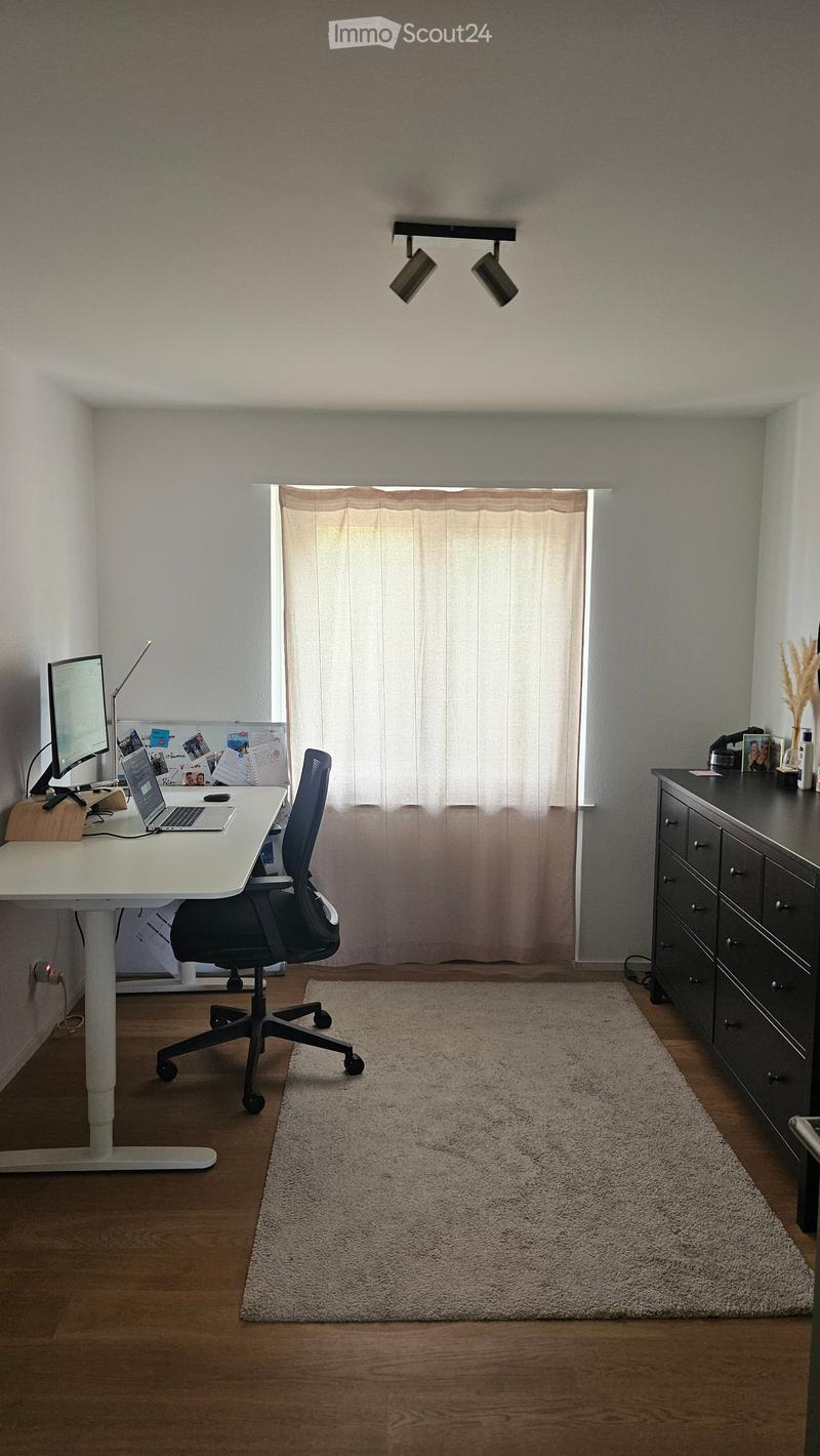 A well-lit room with a wooden floor, a large window with curtains, a desk with a laptop and screen, a black office chair, and a small rug in the center.