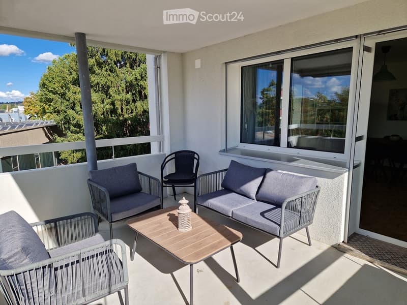 Balcony with a sofa, chairs, a wooden coffee table, and cushions, overlooking a tree-lined area.