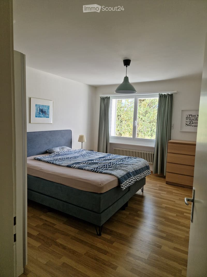Modern bedroom with wooden flooring, large window with grey curtains, grey upholstered bed with patterned bedding, nightstand with lamp, picture frames on walls, and a chest of drawers.