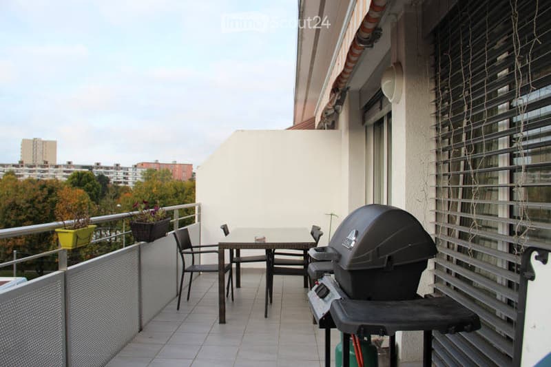 Balcony with a small table, chairs, and a barbecue, overlooking a cityscape with trees and buildings.