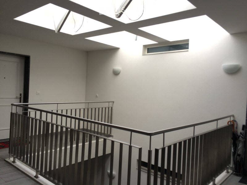 Modern interior hallway with white walls, a metal railing staircase, and skylights providing natural light. The space is clean and minimalistic with a door visible on the left side.