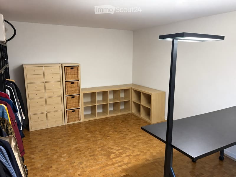 A well-lit room with wooden flooring, a tall wardrobe with multiple drawers, open wooden shelves, a large table with a modern lamp, and a rack of hanging clothes.
