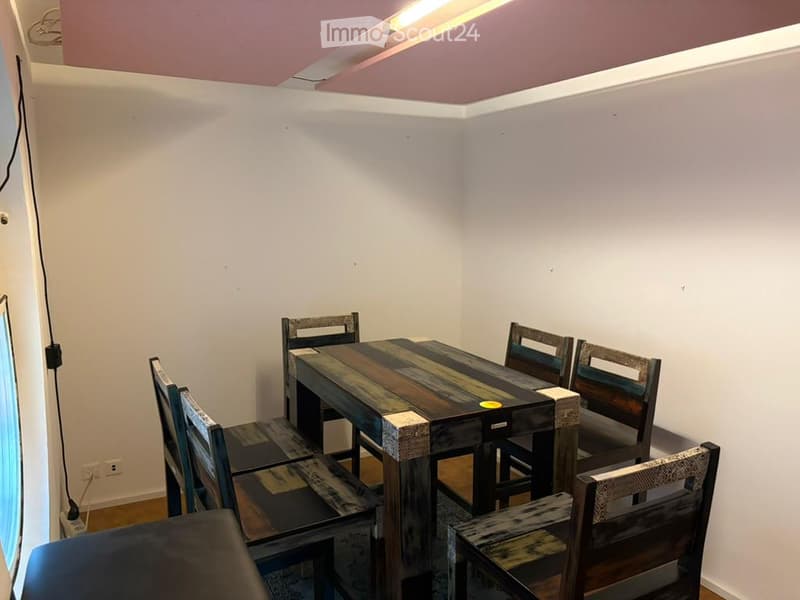 A small, unfurnished room with a wooden table and four matching chairs, white walls, and a window on one side.