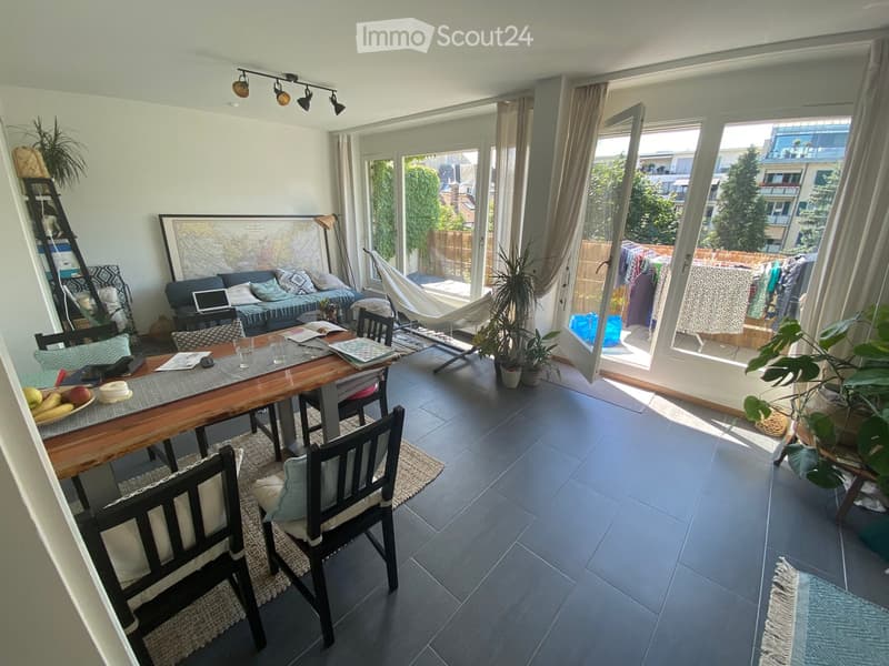 Bright living and dining area with large windows and sliding doors leading to a balcony. Features a sofa, dining table with chairs, laptop, potted plants, and a hammock.