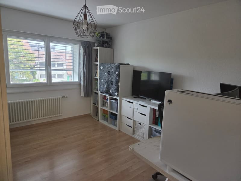 A modern living room with wooden flooring, a large window with a view, a TV on a white cabinet, a shelf with books and decorative items, and a hanging pendant light.