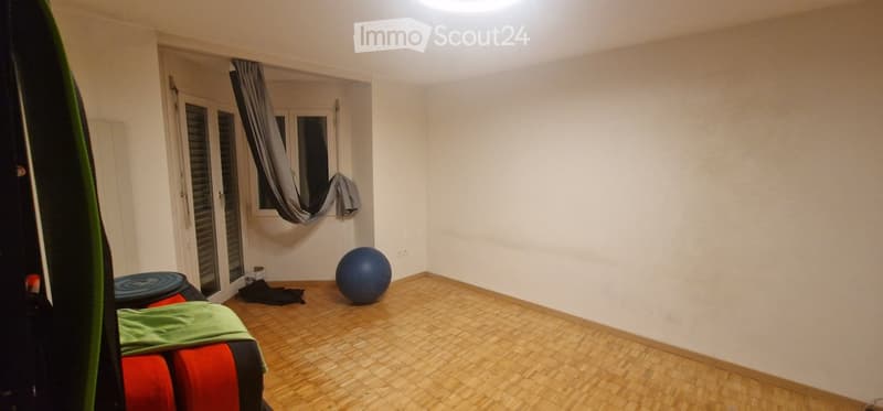 A small, unfurnished room with wooden flooring, a window with a curtain, a mirror, a blue exercise ball, and a red and green cushioned stool.