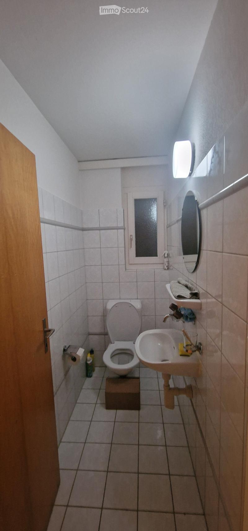 Small bathroom with white tiled walls and floor, featuring a toilet, sink, mirror, and light fixture. Toilet paper and cleaning supplies are visible.