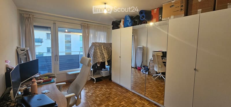 Well-lit room with wooden flooring, large window with balcony access, desk with chair, laptop, and screen, wardrobe with mirror, and various storage items.