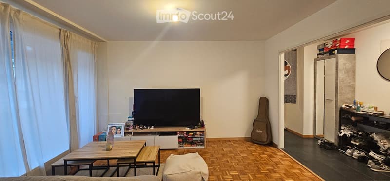 The living room features a wooden floor, a large television on a stand, a wooden coffee table with stools, a guitar case, a mirror, and a shelf with various items.