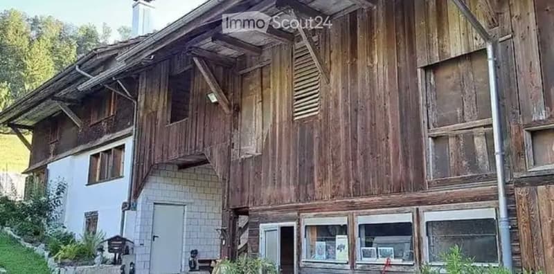 A rustic wooden house with a steeply pitched roof, visible wooden beams, and a mix of wooden and white walls. The house has multiple windows and a small balcony area on the upper level.
