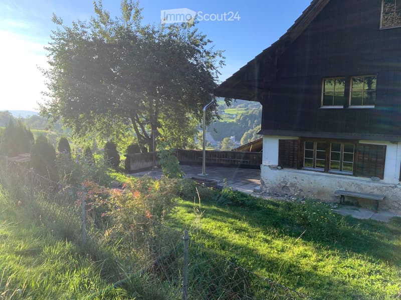 A rustic wooden house with a stone base, surrounded by a well-maintained garden and a wooden deck, with a scenic valley view in the background.