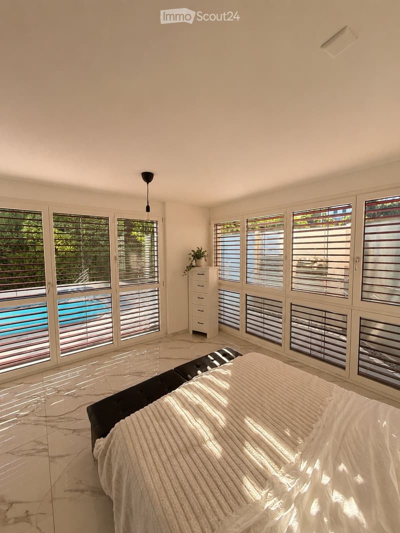 Bright bedroom with large windows, marble flooring, a neatly made bed, a nightstand with a potted plant, and a view of a swimming pool.
